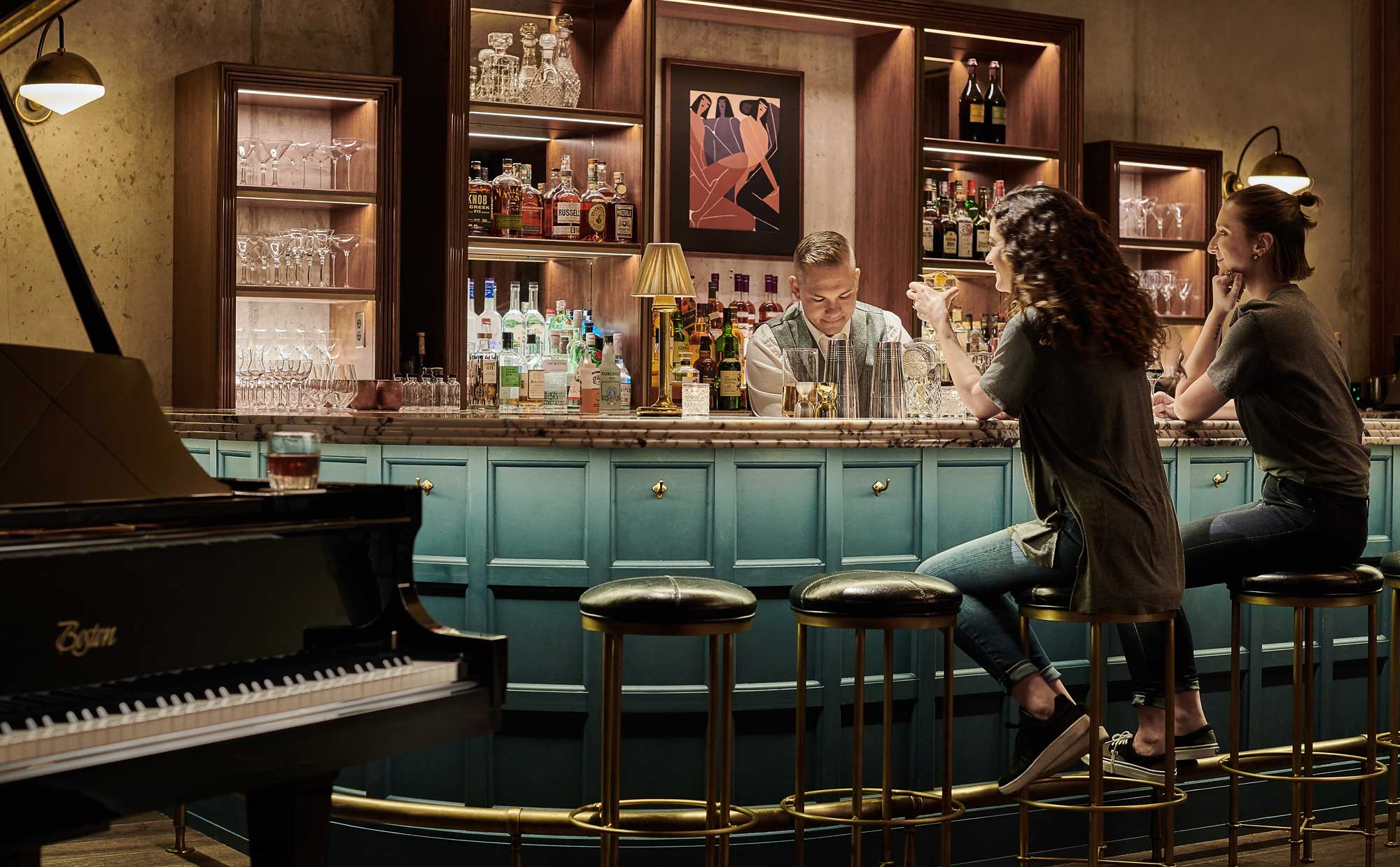 Two women sit at the bar enjoying cocktails in front of a well dressed bar tender, and there is a piano in the foreground