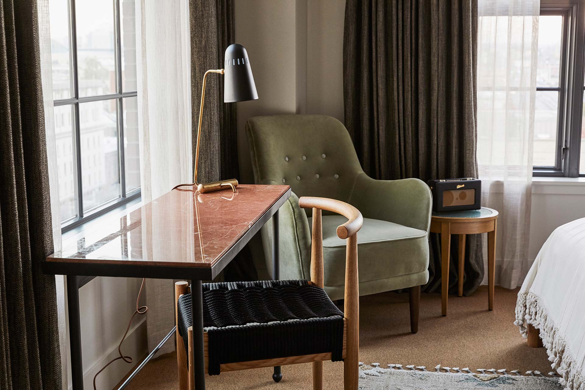 A green lounge chair, small desk and a radio sit in the corner of the hotel room, with windows on both walls