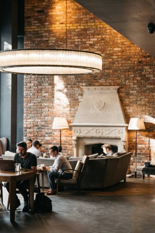 Large hanging circular light over people sitting in the hotel lobby, with an exposed brick wall and fireplace