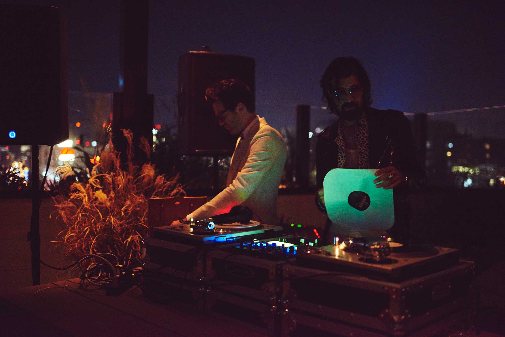 Two DJs at a party on a rooftop with city lights behind