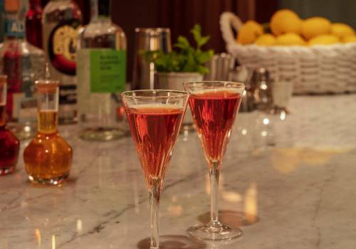 Two drinks sitting in front of spirit bottles and a basket of lemons