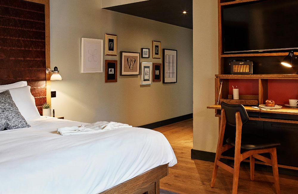 A double bed and desk with TV above it. An assortment of different modern artwork is on the wall leading the door.