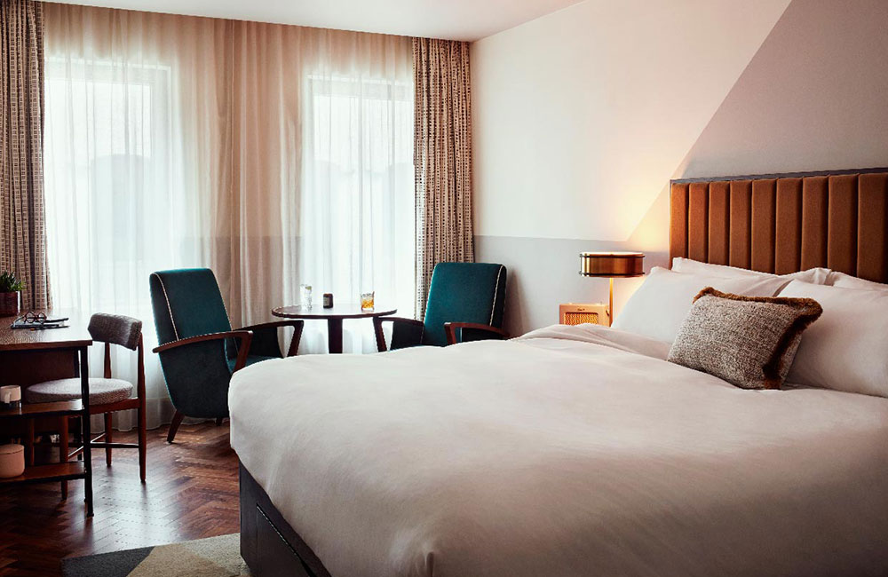 A large double bed, two lounge chairs and table, and a desk area - with two large full length windows in the background.