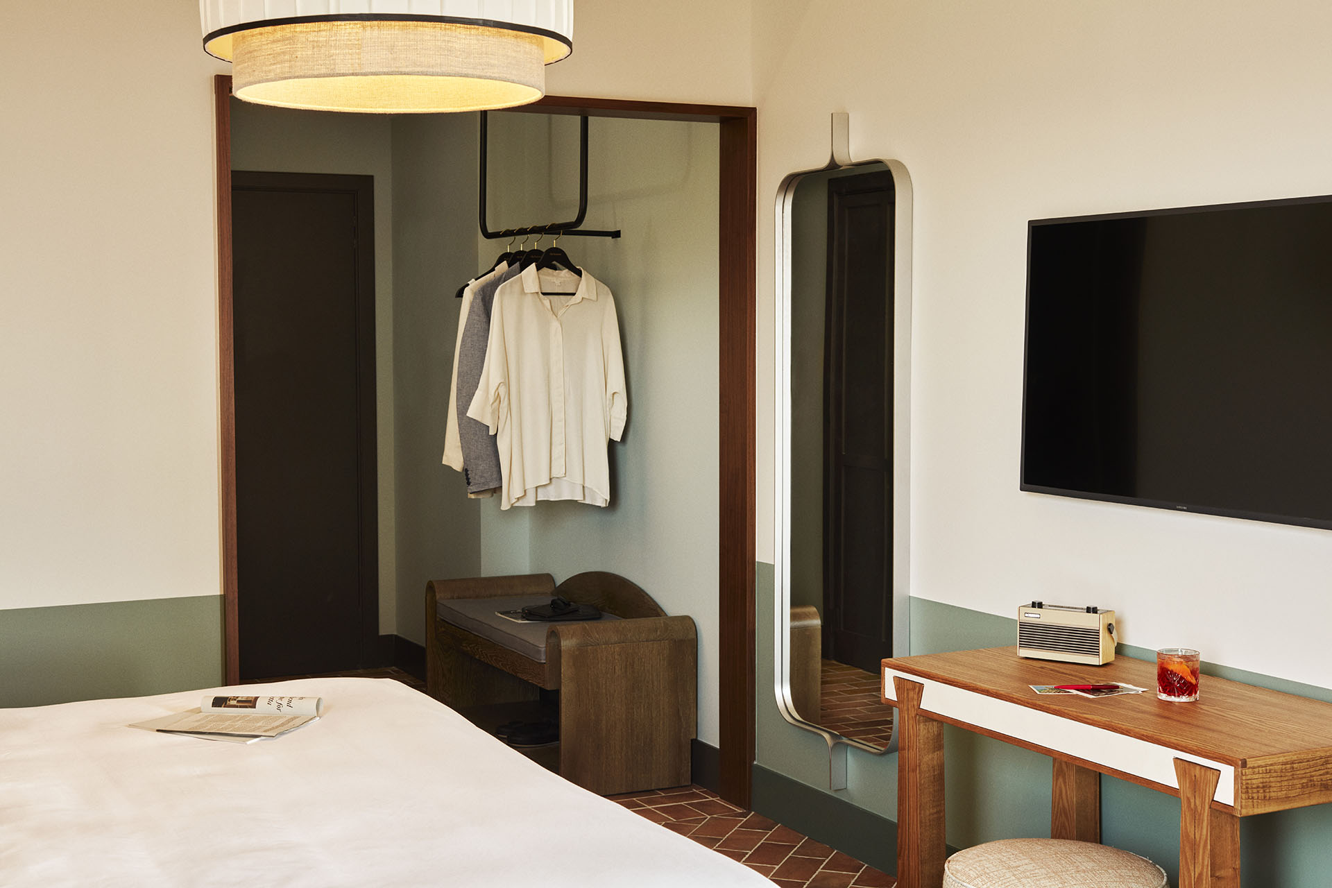 In the room there is a full length mirror on the wall with an area to hang clothes and store luggage. There is a desk underneath the TV