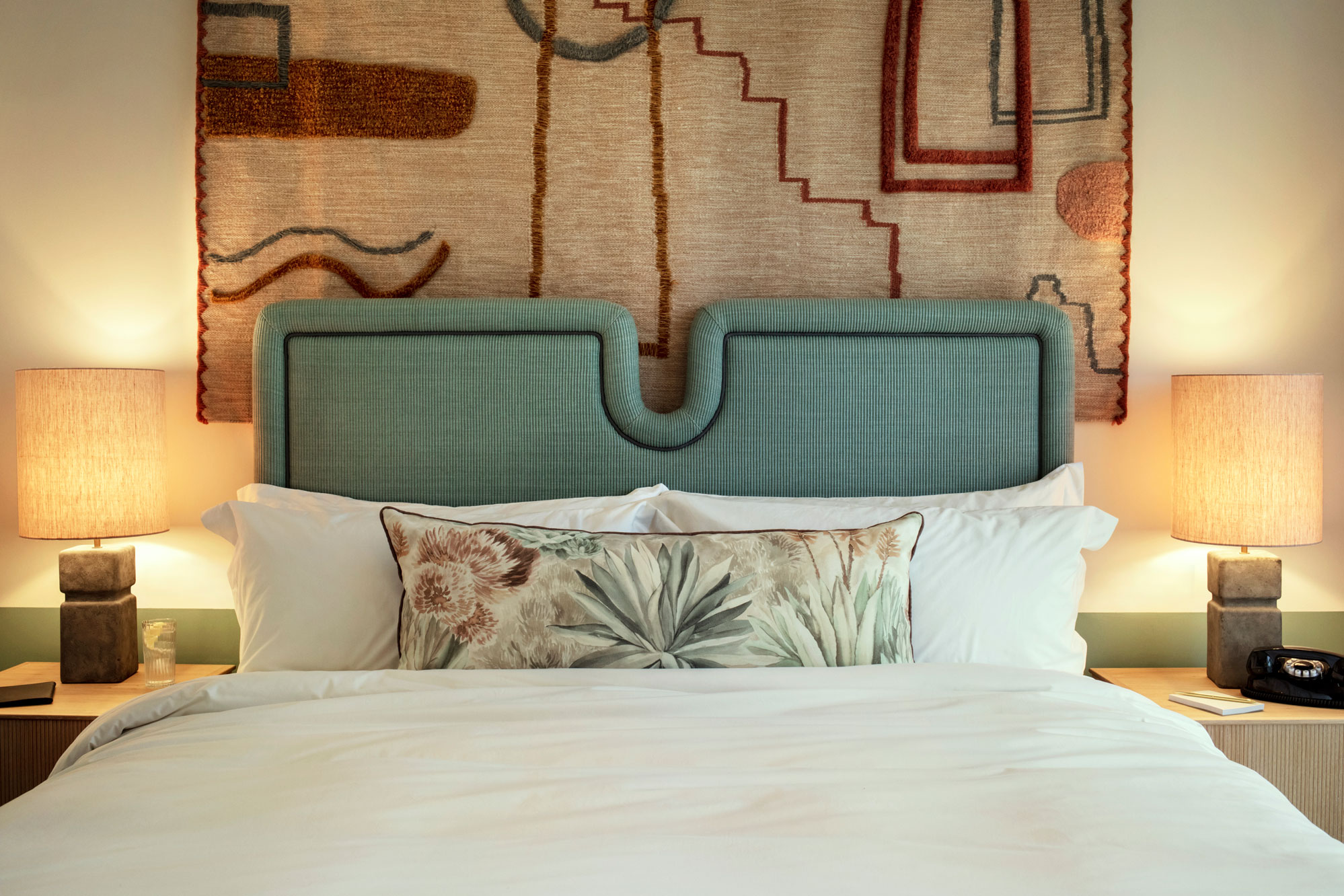 The double bed has side tables with lamps on, a phone and a large decorative hanging above the headboard
