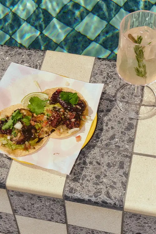 A plate of delicious tacos resting on the side of the pool frame with a tall glass of refreshing beverage with rosemary garnish.