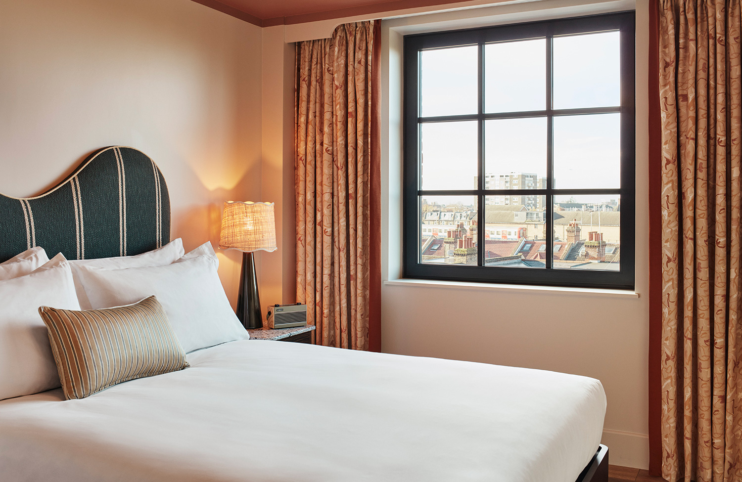 Interior of Cosy room at new hotel in London shows views over Shepherd's Bush, bedside lamp, pastel curtains and comfortable mattress.