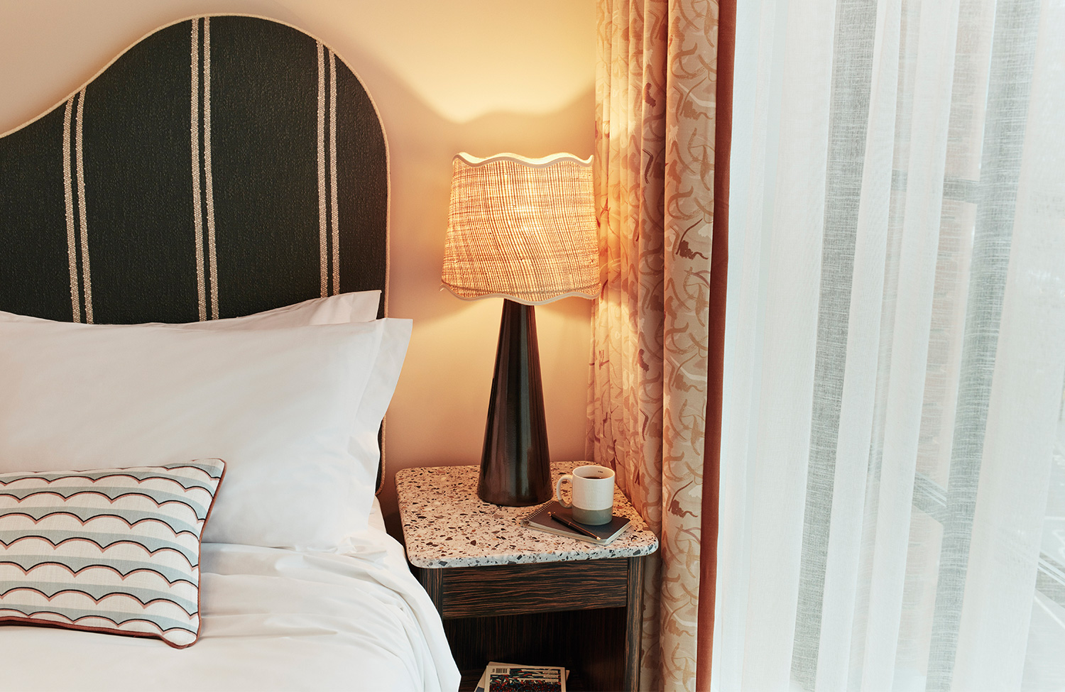 Interior of a Snug room type at The Hoxton, Shepherd's Bush showing soft pillows, bedside lamp and cup of tea or coffee.