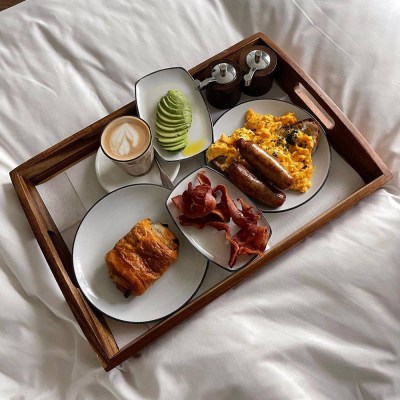A tray of breakfast on a white duvet with avocado, coffee, sausages, scrambled eggs, a pastry and bacon.
