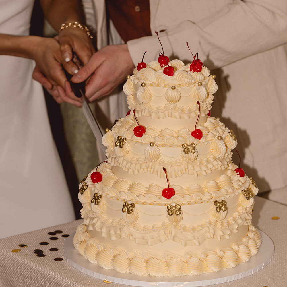 Couple cutting into a white buttercup wedding cake with cherries on the top.