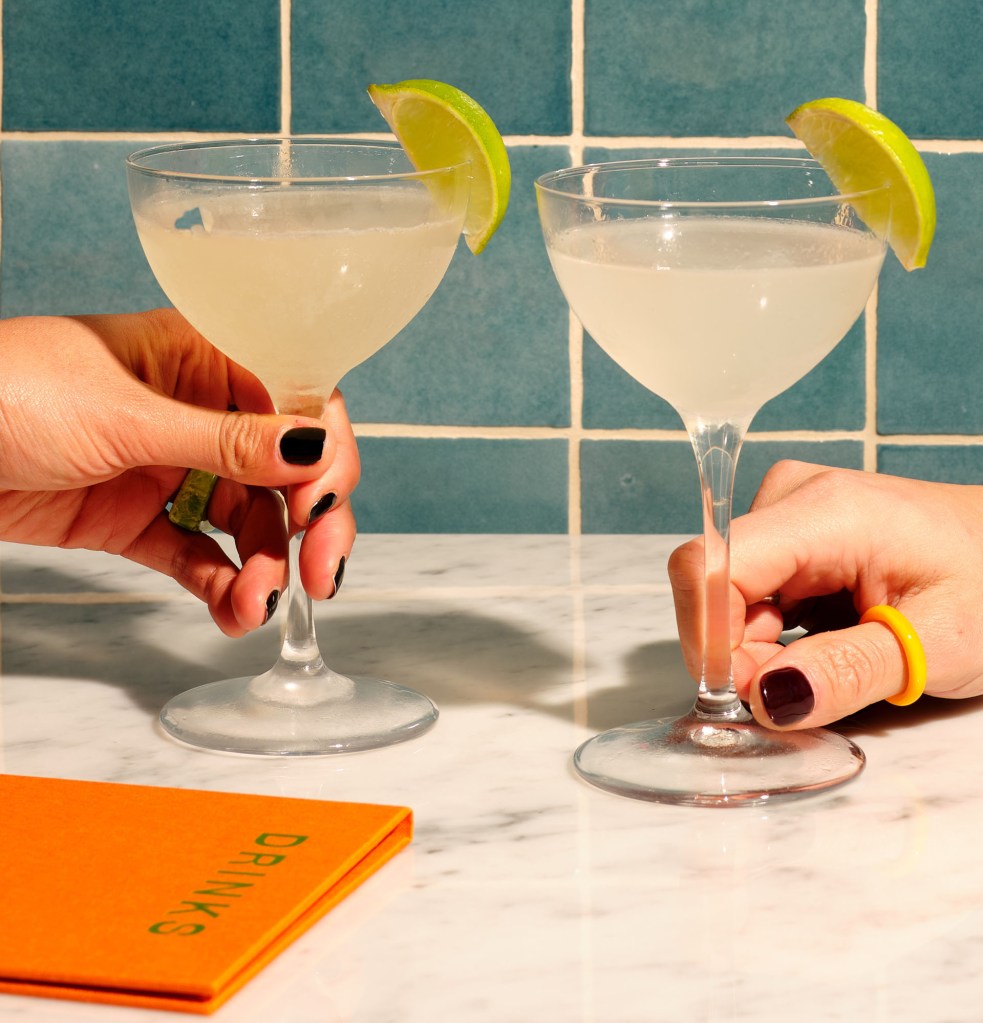 Twee handen die elk een Daiquiri Classico vasthouden, met een menukaart ernaast.