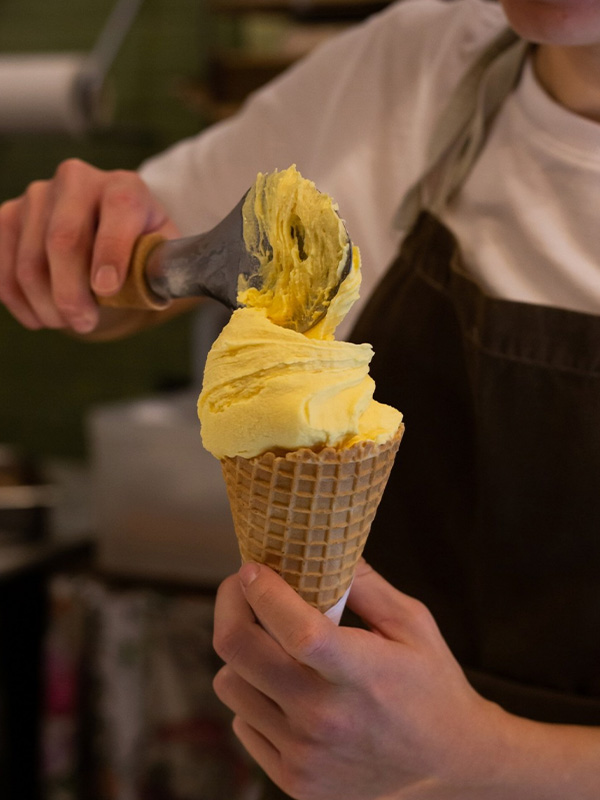 Main tenant un cornet de crème glacée et y versant une cuillère de gelato jaune, chez SBrino.