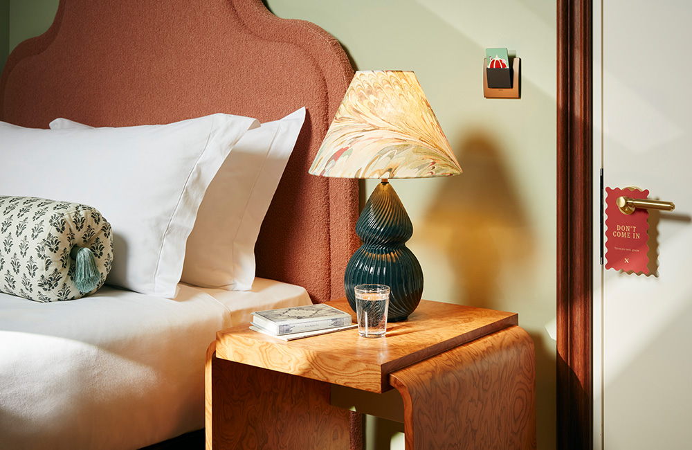Bedside table with a green lamp featuring a marble-patterned lampshade, alongside a bed adorned with large pillows and a decorative bolster cushion.