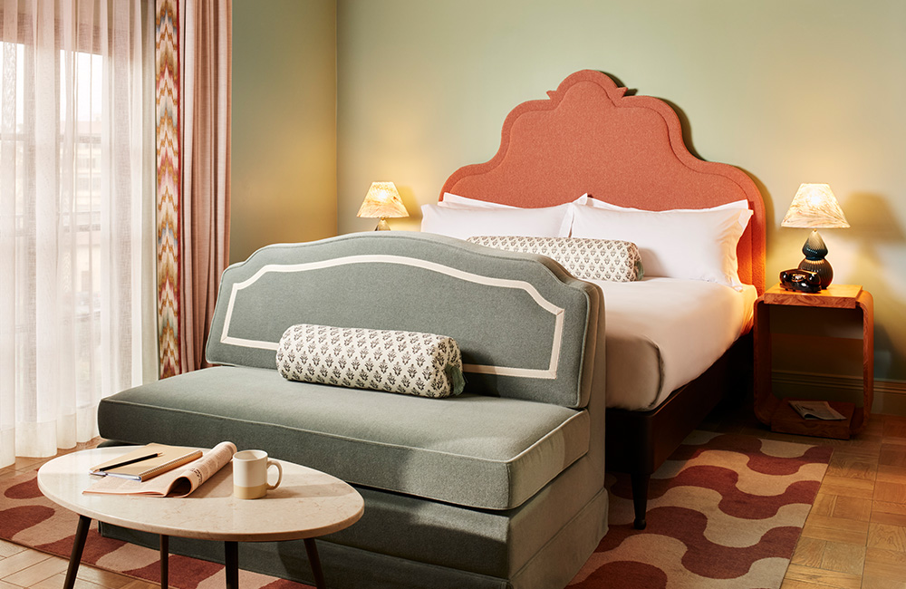 Side view of a soft pastel-colored cozy room, with natural light illuminating a king-size bed. A blue velvet sofa sits at the end of the bed, accompanied by a coffee table.