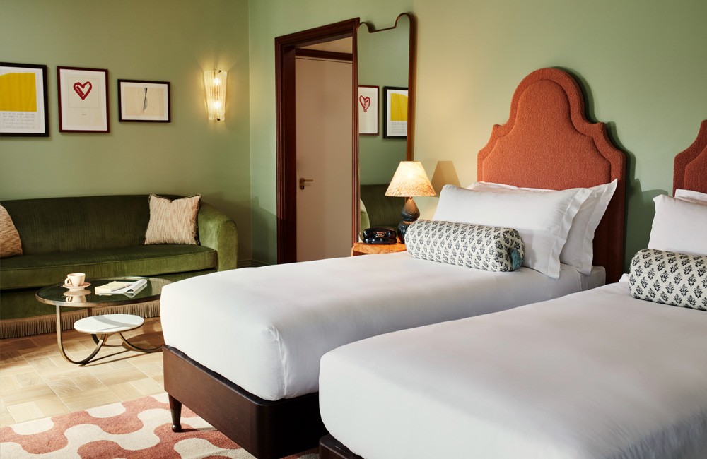 Two single beds with large pastel bolsters on each, accompanied by side lamps on separate bedside tables. A large two-seat sofa and glass coffee table beside the sleeping arrangement.