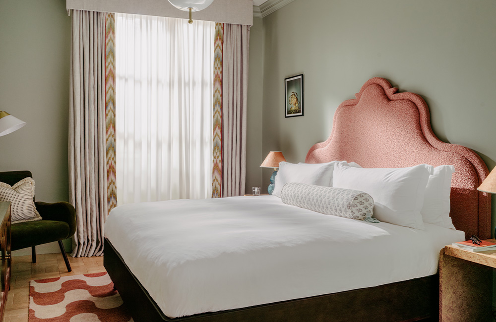 Overview of a spacious hotel room featuring a large king-size bed, an armchair, a bedside table, and a large window allowing natural light to fill the space