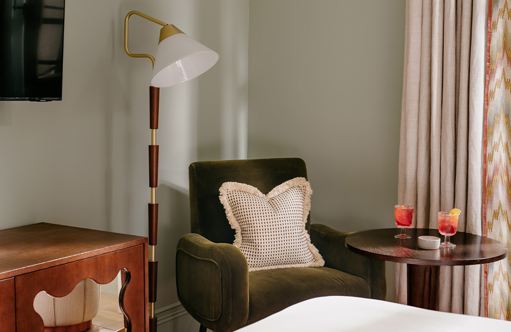 Green velvet armchair by a large window, accompanied by a side table with a bright, refreshing beverage, positioned at the end of the bed.