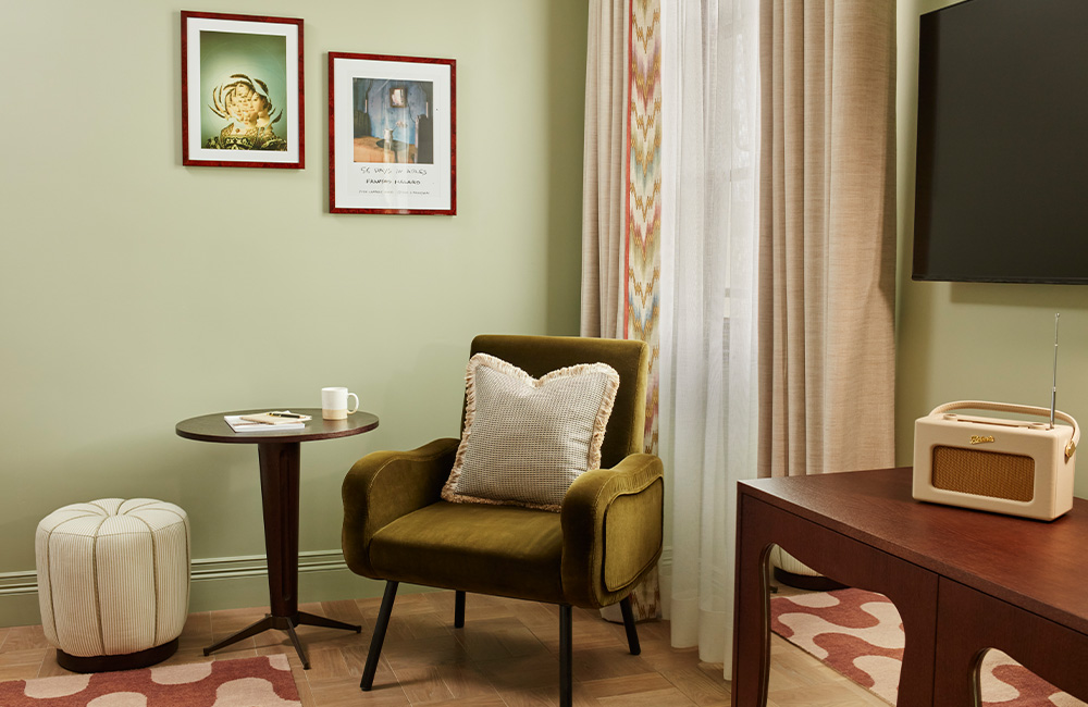 Mint-patterned pouf chair and green velvet armchair with a side table at the end of the bed, positioned near a large window that fills the room with natural light. A large mounted TV with a side table underneath sits beside the window.