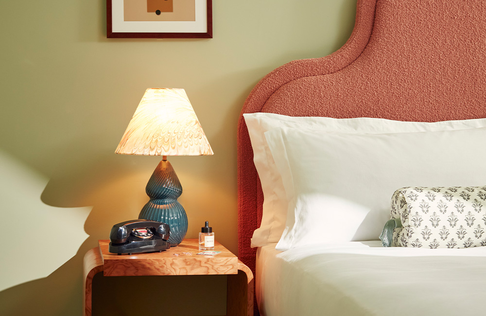 View of a soft pink super king-bed headboard with a bedside table with a green lamp on top.