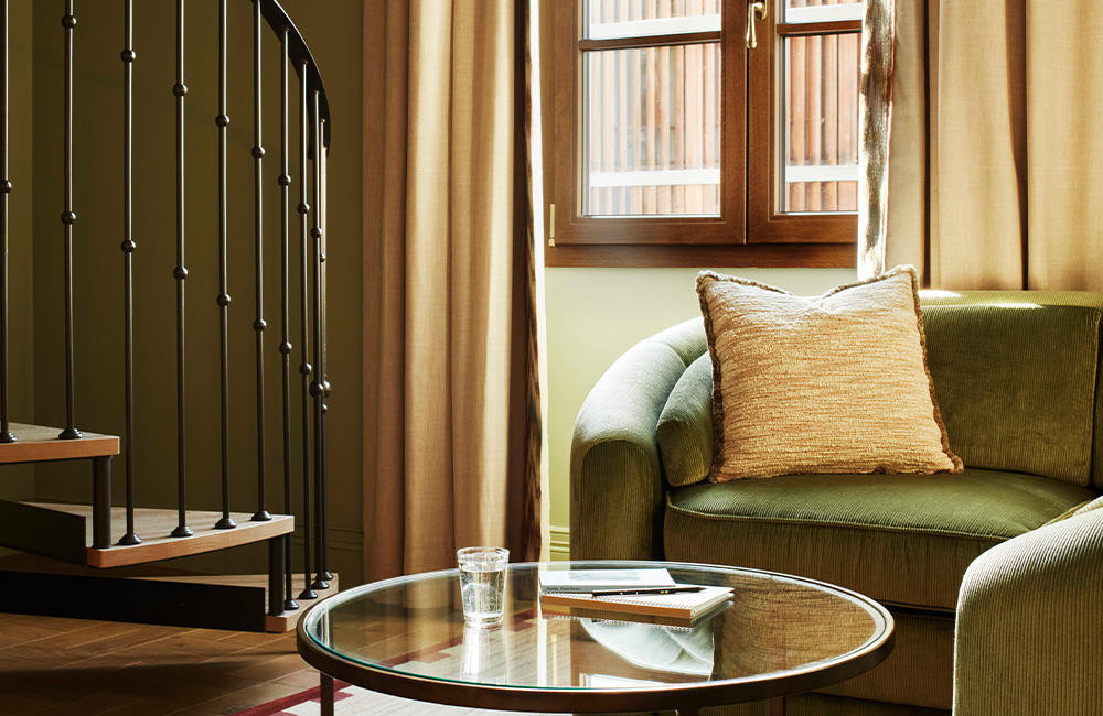Green furniture and a glass coffee table with books, positioned next to a spiral black staircase. A large window fills the space with natural light.