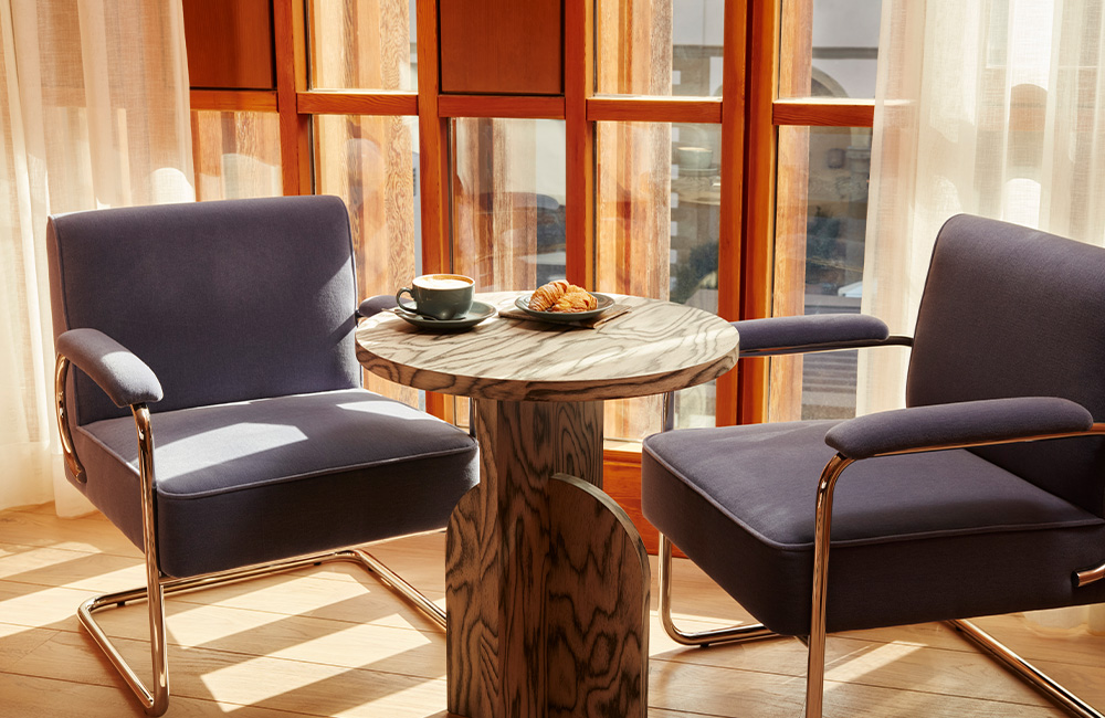 Two blue velvet armchairs with chrome frames, centered around a marble side table holding coffee and pastries. Positioned next to a large window, allowing natural light to fill the hotel room.