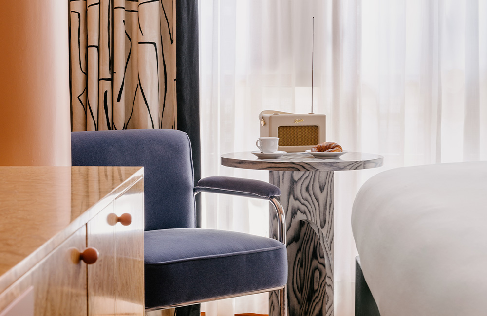End of the bed featuring a blue velvet armchair next to a marble side table, with natural light streaming in from a large window