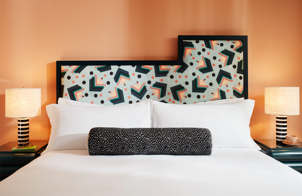 Headboard with a bright, dynamic pattern, large fluffy pillows, and a black and white lamp beside on both side of the bed.