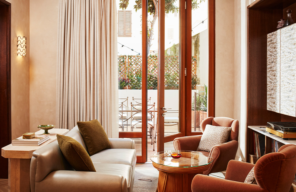 Seating area with s suede leather sofa, a console marble table against the back of the sofas, and two soft velvet armchairs facing a coffee table. Large terrace doors are located behind the seating cluster.
