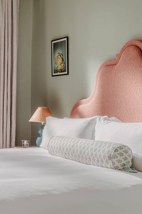 Close-up side view of a pink headboard with large white pillows and a soft-patterned bolster cushion, with natural light coming through a large window.